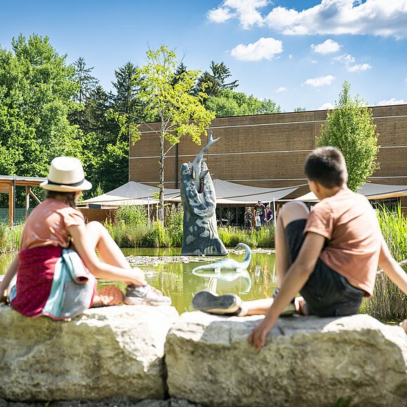 Dinosauriermuseum in Denkendorf Zwei Kinder sitzen am Wasser im Außengelände des Dinosauriermuseum und beobachten die grüne Landschaft und das Wasser.
